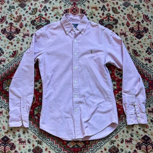 Rosa skjorta ifrån raulph lauren - Rosa skjorta från raulph. Slim fit. Säljer för den är för liten för mig. Mått: längd 73 cm, axelbredd 44 cm, bröstbredd 50 cm, armlängd ifrån armhålan 51 cm. Liten defekt på magen annars väldigt bra skick 