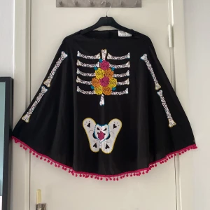 Svart skelett-tröja med rosor och silkes handskar med glasknappar - Unik svart tröja med färgglatt skelett-tryck och rosor i gult, orange och rosa på bröstet. Vida ärmar och dekorativ kant med röda bollar. Perfekt för halloween eller temafest. Materialet känns mjukt och lätt, troligen polyester.