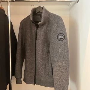 Grå fleecekofta från Canada Goose - Säljer en grå fleecekofta från Canada Goose med broderad logga på ärmen. Koftan har ribbade muddar, hög krage och hel dragkedja framtill. Materialet är mjukt och värmande, perfekt för kyliga dagar. Kvitto och andra bevis på produktens äkthet tillgängliga. Storlek S, men passar även bra för M 