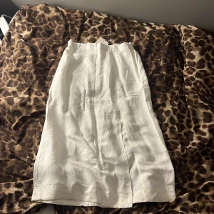 Vit långkjol i linne från GANT - Snygg vit långkjol från GANT i mjukt linne. Kjolen har en rak siluett med slits framtill som ger extra rörelsefrihet. Midjan är elastisk för skön passform och stilen är clean och fräsch, perfekt för dig som gillar minimalistiska vibes. Oanvänd!! Prislappen kvar på.