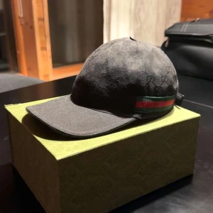 Svart Gucci keps med GG-mönster - Snygg svart keps från Gucci med diskret GG-monogram över hela ytan. Klassisk böjd skärm och justerbar rem bak. På sidan finns Guccis ikoniska röd-gröna band. Materialet är bomull och kepsen har en stilren, modern look som passar till streetwear.