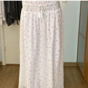 Vit långkjol med blommigt mönster - Supersöt vit långkjol med små rosa blommor och gröna detaljer. Kjolen har en smockad midja med snörning och tofsar som ger en chill och romantisk vibe. Perfect för varma dagar. ⚠️Inte mina bilder⚠️