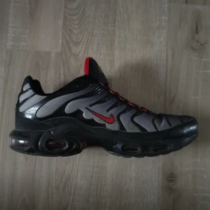 Nike Air Max Plus i svart och grå - Snygga Nike Air Max Plus sneakers i svart och grå med röda detaljer. Skorna har en unik design med vågformade linjer och en synlig Air Max-dämpning i sulan för extra komfort. Perfekta för både stil och funktion.