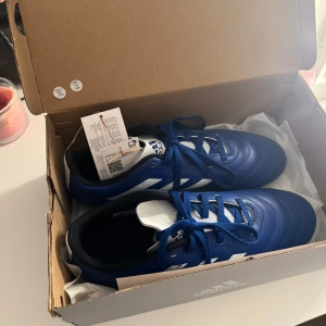 Blå fotbollsskor från Adidas - Snygga blå fotbollsskor från Adidas med vita detaljer och klassisk snörning. Perfekta för fotbollsplanen med en stilren design och bekväm passform.