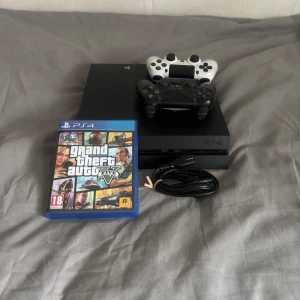 Grand Theft Auto V till PS4 - Säljer Grand Theft Auto V till PS4. Spelet kommer i originalförpackning med omslag i färgglada illustrationer. Perfekt för dig som älskar action och äventyr i en öppen värld. Inkluderar även en PS4-konsol med två handkontroller, en svart och en vit, samt en strömkabel.