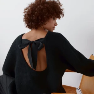 Svart stickad tröja med rosett - Säljer en elegant svart stickad tröja med en öppen rygg och en dekorativ rosett. Tröjan har långa ärmar och en mjuk, bekväm passform. Perfekt för en stilren look.