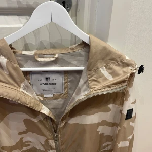 Beige och vit Camo Windbreaker från Woolrich - Unik Beige/Vit Camo Windbreaker från Woolrich. Storlek: L. Skick: 9/10, väldigt fint skick! Kontakta oss för fler bilder eller info!🙌