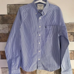 Blå och vit randig skjorta från Abercrombie & Fitch - Säljer en stilren blå och vit randig skjorta från Abercrombie & Fitch. Skjortan har långa ärmar och en klassisk krage med knappar framtill. Perfekt för en avslappnad men ändå uppklädd look.