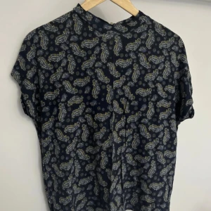 Mörkblå blus med fjäder mönster Medium - Mörkblå blus med vitt och gult mönster som fjädrar. Öppning i nacken och Loose fit. Storlek Medium från Holly & Whyte by Lindex