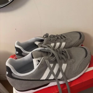 Grå Adidas sneakers - Snygga grå sneakers från Adidas med klassiska vita ränder på sidan. Skorna har snörning och en bekväm passform, perfekt för vardagsbruk. De är tillverkade i en kombination av mocka och syntetmaterial för en stilren look.