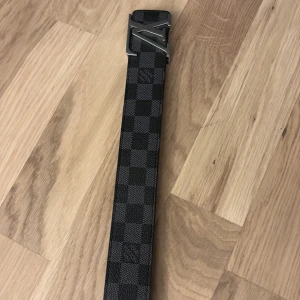 Rutmönstrat bälte med metallspänne Louis vuttion - Snyggt bälte med ett klassiskt rutmönster i svart och grått. Bältet har ett stilrent metallspänne som ger en elegant touch. Perfekt för att lyfta vilken outfit som helst. Obs har ett Exstra hål