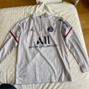 Grå långärmad träningströja från Jordan - Säljer en grå långärmad träningströja från Jordan med PSG-logga och texten 'Accor Live Limitless' på framsidan. Tröjan har en halv dragkedja och prickigt mönster. Perfekt för fotbollsträning eller annan sportaktivitet.