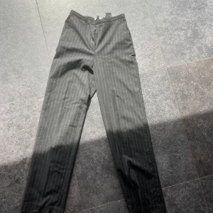 Svarta randiga kostymbyxor - Snygga svarta kostymbyxor från H&M med diskreta vita ränder. Byxorna har en klassisk passform och är perfekta för en stilren look. De har en dragkedja framtill och är tillverkade i ett bekvämt material.