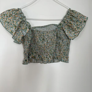 Blommig offshoulder topp - Söt blommig offshoulder topp i ljusgrönt med små gula och vita blommor. Toppen har en smockad design och volangdetaljer vid ärmarna och nederkanten. Lite använd och är i bra skick. Hittar inga defekter på den. 