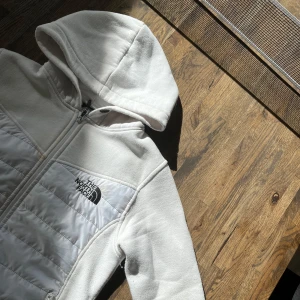 Vit cardigan jacka från The North Face - Säljer en vit fleecejacka från The North Face med huva och dragkedja. Jackan har quiltade paneler på fram- och baksidan samt två fickor med dragkedja. Perfekt nu till våren!