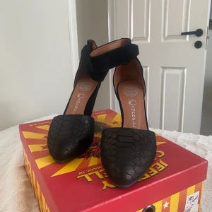 Svarta klackar från Jeffrey Campbell - Snygga svarta klackar från Jeffrey Campbell med en unik textur på framsidan. Skorna har en rund tå och en bred ankelrem för extra stöd. Perfekta för att ge lite höjd och stil till din outfit. Skorna är aldrig använda. Orginalpris 1300