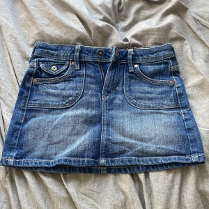 Blå jeanskjol från NOW - Säljer en snygg blå jeanskjol från NOW med klassisk femficksdesign och dekorativa knappar på bakfickorna. Kjolen har en kort längd och en stilren look som passar perfekt till vardags. Den har en blommig insida i midjan som ger en extra touch av stil.