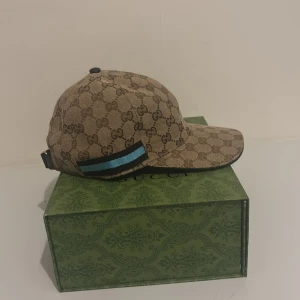 Beige keps från Gucci Limited edition  - Snygg beige keps från Gucci med det klassiska GG-mönstret. Kepsen har en justerbar rem baktill och en blå och svart randdetalj på sidan. Perfekt för att ge din outfit en lyxig touch.