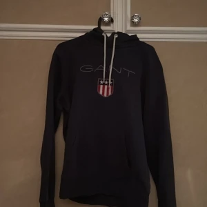Mörkblå hoodie från GANT - Säljer en snygg mörkblå hoodie från GANT med deras ikoniska logga och ett sköldmärke på bröstet. Tröjan har en justerbar huva med vita snören och en praktisk magficka. Perfekt för en avslappnad stil.