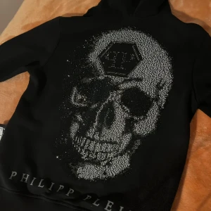 Äkta svart hoodie med dödskalle från Philipp Plein - Snygg svart hoodie från Philipp Plein med en stor dödskalle i glittrande detaljer på ryggen. Tröjan har dragkedja framtill och en klassisk huva. Perfekt för en cool och edgy stil.