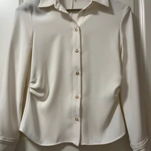 Vit blus från Zara  - Elegant vit blus med långa ärmar och knappar framtill. Blusen har en klassisk krage och dekorativa knappar vid ärmsluten. Perfekt för en stilren look.