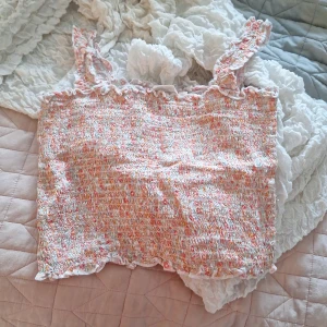 Topp - Söt blommig topp från KappAhl i en mix av rosa, orange och vit. Toppen har en smockad design med volangdetaljer vid axelbanden och nederkanten. Perfekt för varma sommardagar!