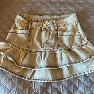 Grå jeans kjol - Trendig beige jeanskjol med volangdetaljer och knapp framtill. Perfekt för en avslappnad stil med bälteshällor för extra stylingmöjligheter.