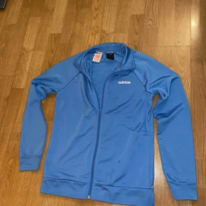 Blå träningsjacka från Adidas - Säljer en blå träningsjacka från Adidas med dragkedja och långärmade ärmar. Jackan har en enkel design med Adidas-loggan på bröstet. Perfekt för träning eller som en lätt överdragströja.