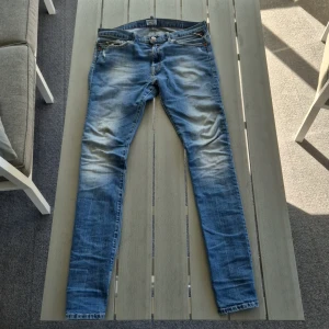 Blå jeans från Replay - Snygga blå jeans från Replay med en klassisk femficksdesign och lätt slitna detaljer. Jeansen har en knappgylf och är tillverkade i ett bekvämt denimtyg. Perfekta för en avslappnad stil. Liten fläck pp knär men det går bort i tvätten