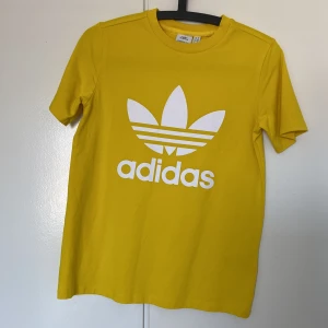 Gul t-shirt Adidas Originals - Säljer en gul t-shirt från Adidas med det klassiska trefoil-logotypen i vitt på framsidan. Perfekt för en sportig och avslappnad stil. T-shirten är kortärmad och gjord i ett mjukt material.