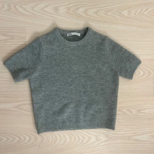 Grå stickad tröja från Zara - Säljer en stilren grå stickad tröja från Zara. Tröjan har korta ärmar och en rund halsringning, perfekt för lager-på-lager eller som den är. Den är mjuk och bekväm, idealisk för både vardag och avslappnade tillfällen.