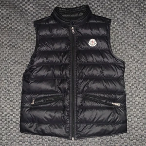 Svart dunväst från Moncler - Säljer en stilren svart dunväst från Moncler med dragkedja och två praktiska fickor framtill. Västen har en quiltad design och Monclers logo på bröstet. Helt äkta, passar 12-14 åringar.