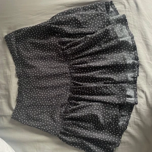 Svart prickig kjol - Säljer en söt svart kortkjol med vita prickar. Kjolen har en volangdetalj som ger den extra charm och rörelse. Perfekt för en dag ute eller en kväll med vänner. Passar bra med en enkel topp eller blus.