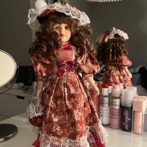 Vintage docka i klänning med spetsdetaljer - Charmig vintage docka med lockigt brunt hår och en vacker klänning i rödbrunt med blommönster. Klänningen har spetsdetaljer och en matchande hatt. 💕Pris kan diskuteras💕
