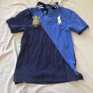 Blå och marinblå pikétröja från Ralph Lauren - Säljer en snygg pikétröja från Ralph Lauren i blå och marinblå färg. Tröjan har korta ärmar och en klassisk krage med ränder. Den har broderade detaljer på bröstet och knappar framtill. Perfekt för en stilren look! L i barnstorlek vilket motsvarar XS/S