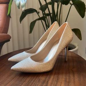 Glittriga guldiga pumps - Snygga glittriga pumps i guld med spetsig tå och mellanhög klack. Perfekta för att ge en glamorös touch till din outfit. Passar utmärkt till festliga tillfällen. Riktiga kvalitet klackar.