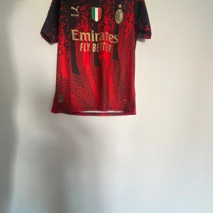 Röd och svart fotbollströja från Puma - Säljer en snygg röd och svart fotbollströja från Puma med korta ärmar. Tröjan har ett coolt mönster och texten 'Emirates Fly Better' på framsidan. På baksidan finns namnet och numret 'Ibrahimovic 11'. Perfekt för fotbollsfans!