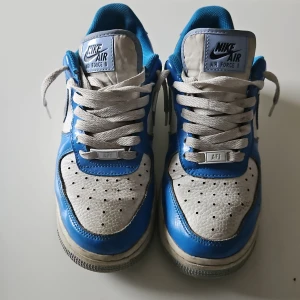 Nike Air Force 1 i blått och vitt - Säljer ett par klassiska Nike Air Force 1 sneakers i en fräsch blå och vit färgkombination. Skorna har en perforerad tå och vit snörning. Perfekta för en stilren look.