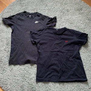 2-Pack T-Shirts  - Två svarta t-shirts till salu. Den ena är från Nike med ett vitt broderat logotyp på bröstet. Den andra är från Fred Perry med ett rött broderat emblem. Perfekta för en casual look! Nypris för båda: 800 kr