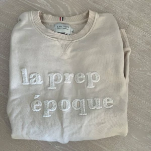Beige sweatshirt från Les Deux - Säljer en stilren beige sweatshirt från Les Deux med texten 'la prep époque' broderad på framsidan. Tröjan har en rund halsringning och ribbade muddar vid ärmslut och nederkant. Perfekt för en avslappnad stil. Strl L. Som ny! Rök och djurfritt hem.