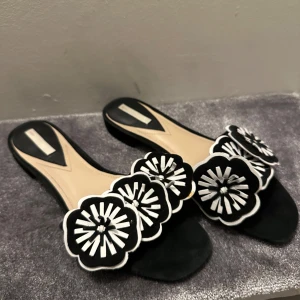 Svarta sandaler med blommig detalj - Svarta sandaler med stora vita blommor. De har en öppen tå och en låg klack. 