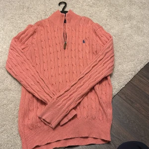 Korallfärgad stickad tröja från Ralph Lauren - Säljer en snygg korallfärgad stickad tröja från Ralph Lauren med kabelstickat mönster och halv dragkedja. Perfekt för kyliga dagar med sin mjuka och bekväma passform. Tröjan har långa ärmar och ribbade kanter. Passa på att fynda denna stilrena klassiker! 🍂