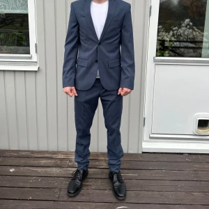 Mörkblå kostym - Snygg mörkblå kostym med kavaj och matchande byxor. Kavajen har en enkelknäppning med två knappar och klassiska slag. Byxorna har en rak passform. Perfekt för formella tillfällen.