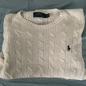 Beige stickad tröja från Ralph Lauren - Säljer en klassisk beige stickad tröja från Ralph Lauren med kabelstickat mönster. Tröjan har långa ärmar och en rund halsringning. Den ikoniska loggan är broderad på bröstet. Perfekt för en stilren och tidlös look. Tröjan är i helt nytt skick. Det är storlek S. Fråga vid minsta lilla fundering!