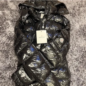 Svart dunjacka moncler - Snygg svart dunjacka från med glansig finish och huva. Jackan har en dragkedja framtill och långa ärmar. Perfekt för kyliga dagar. Prislappen är borta tyvärr. Storlek S men sitter stort på så skulle säga som en Medium