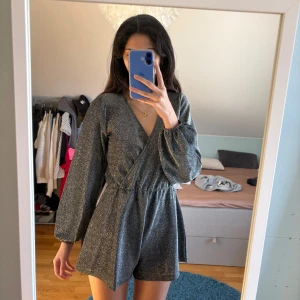 Glittrig byxdress/jumpsuit - (Ej klänning!) Säljer denna fina korta glittriga byxdressen/jumpsuiten. Den har långa ärmar och en elastisk midja för en bekväm passform. Kan även bäras off shoulder. Endast använd en gång. Perfekt för nyår eller glitterfest!!