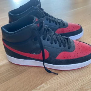 Nike sneakers i svart och rött - Snygga Nike sneakers i svart och rött med hög design och klassisk snörning. Skorna har en vit sula och det ikoniska Nike-märket i rött på sidan. Perfekta för en sportig look. Säljer dem då de var lite stora och inte min stil, kartong saknas. 