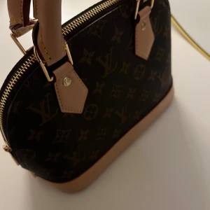 Alma bb handväska från Louis Vuitton - Elegant handväska i klassiskt monogrammönster från Louis Vuitton. Väskan har en dragkedja upptill och detaljer i ljusbrunt skinn. Perfekt för att ge en lyxig touch till din outfit.