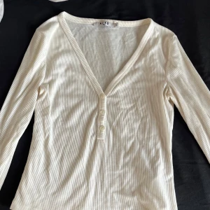 Vit ribbad tröja från NA-KD - Säljer en stilren vit ribbad tröja från NA-KD med v-ringning och knappar framtill. Perfekt för en avslappnad look. Tröjan har långa ärmar och är tillverkad i ett mjukt material.