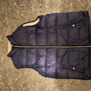 Vändbar dunväst i skinigt blått och grått - Säljer en snygg vändbar dunväst med en sida i marinblått och den andra i grått. Köpt från ralph lauren hemsidan när de fortfarande fanns. Västen sitter stilrent men har bara en mindre defekt på den blåa sidans ficka som man kan se på bild 5. Riktigt fin väst som kan passa båda XS och S. Säljer eftersom att jag köpt en ny väst. Priset kan även diskuteras.